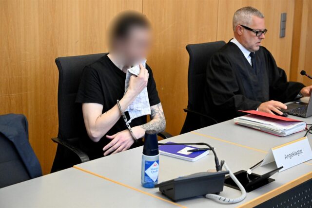 Der Angeklagte sitzt im Landgericht in Ulm. Er hält sich ein Spültuch vor dem Mund, weil er durch die Schüsse eines Polizisten am Kiefer verletzt wurde und nun nicht mehr richtig trinken kann.