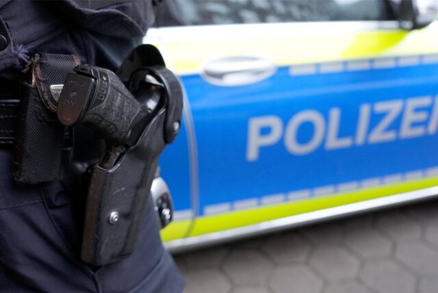 Die Dienstwaffe eines Polizeibeamten, der vor einem Streifenwagen steht.