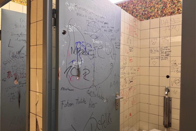 Ein Blick in die Schul-Toiletten der Willy-Brandt-Gesamtschule Bergkamen. Alles, also Wandkacheln, Tür und Wand sind mit Kritzeleien überzogen, die Tür ist beschädigt, Toilette und Spülung uralt.