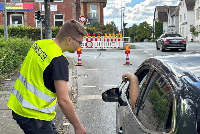 Ein Mann mit einer gelben Weste steht in Unna an der Ecke Beethovenring/Obere Husemannstraße. Ein Autofahrer zeigt ihm einen Ausweis.