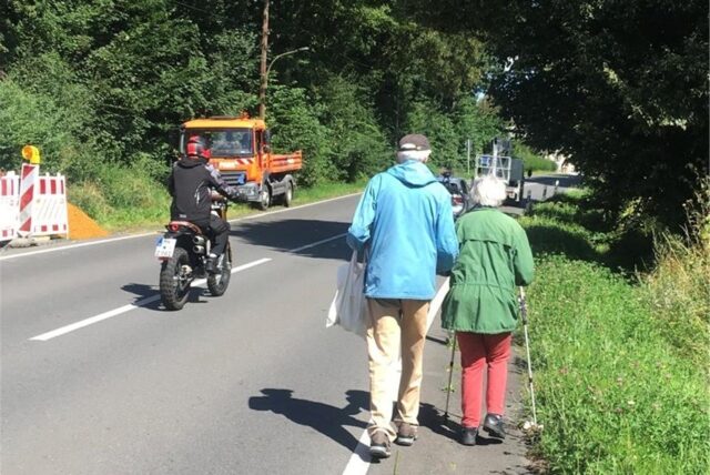 Zwei ältere Menschen gehen in Holzwickede-Hengsen an der Schwerter Straße entlang. Zu erkennen ist, dass ein Mann fast auf der Straße laufen muss. Das Unfallrisiko ist entsprechend hoch.