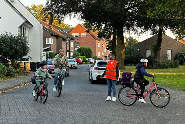 Eine Frau steht mir einer Warnweste bekleidet auf der Straße und achtet darauf, dass Fahrradfahrer sicher abbiegen können.