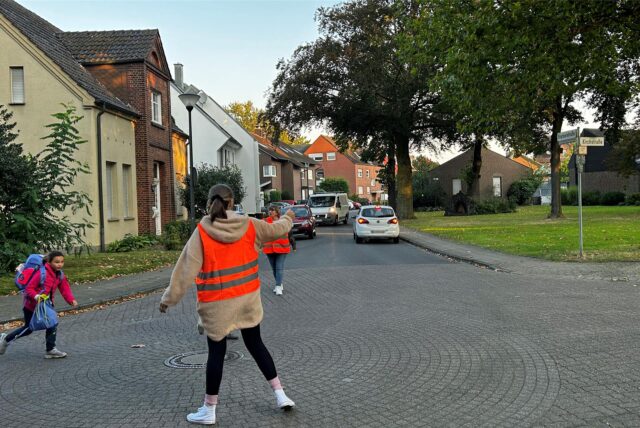 Frauen stehen in Warnwesten auf einer Kreuzung und helfen so einem Kind über die Straße.