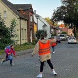 Frauen stehen in Warnwesten auf einer Kreuzung und helfen so einem Kind über die Straße.