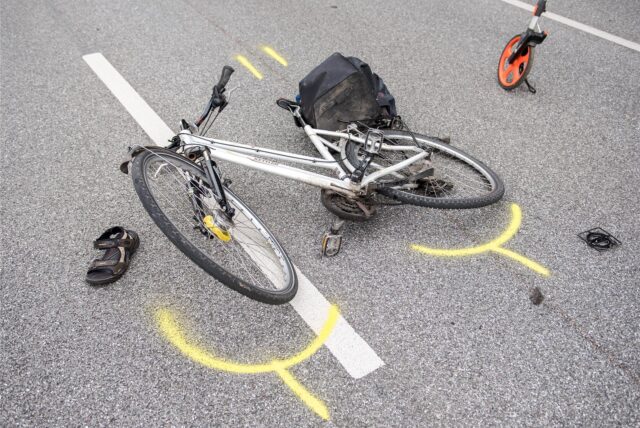 Ein weißes Fahrrad liegt verbeult auf einer Straße. Es sind gelbe Markierungen zu sehen.