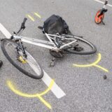 Ein weißes Fahrrad liegt verbeult auf einer Straße. Es sind gelbe Markierungen zu sehen.