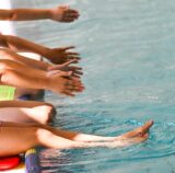 Aus dem Bildrand heraus schauen Kinderhände und -füße, von Kindenr, die am Beckenrand eines Schwimmbeckens sitzen.
