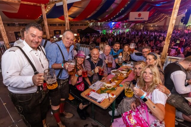Elf Besucher in Oktoberfest-Outfits sitzen an einem länglichen Tisch, posieren für die Kamera und halten Bierkrüge hoch. Im Hintergrund sieht man eine große Menge weiterer Besucher im Festzelt.