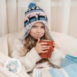 Mädchen mit Pudelmütze und Winterdecke auf einem Sessel, eine große rote Tasse in der Hand.