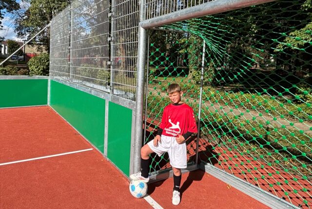 Ein Schüler der Fußball-AG der Schule am Friedrichsborn steht im Tor auf dem wiedereröffneten Bolzplatz an der Eiswiese im Kurpark. Er hat ein rotes Trikot an und eine weiße Trikothose und stützt einen Fuß auf einen Fußball.