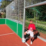 Ein Schüler der Fußball-AG der Schule am Friedrichsborn steht im Tor auf dem wiedereröffneten Bolzplatz an der Eiswiese im Kurpark. Er hat ein rotes Trikot an und eine weiße Trikothose und stützt einen Fuß auf einen Fußball.