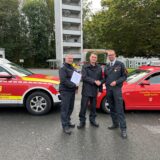 Dirk Kemke (Leiter der Feuerwehr Bergkamen) und Volker Rost (Leiter der Feuerwehr Kamen) stehen mit Tim Krächter vor Feuerwehrfahrzeugen der beiden Städte an der Feuer- und Rettungswache im Mersch und überreichen ihm die Ernennungsurkunde.