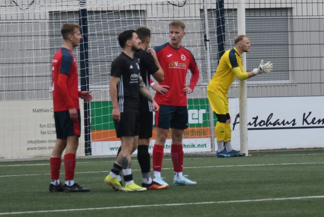 Fußballer in schwarzen und roten Trikots stehen im Strafraum.