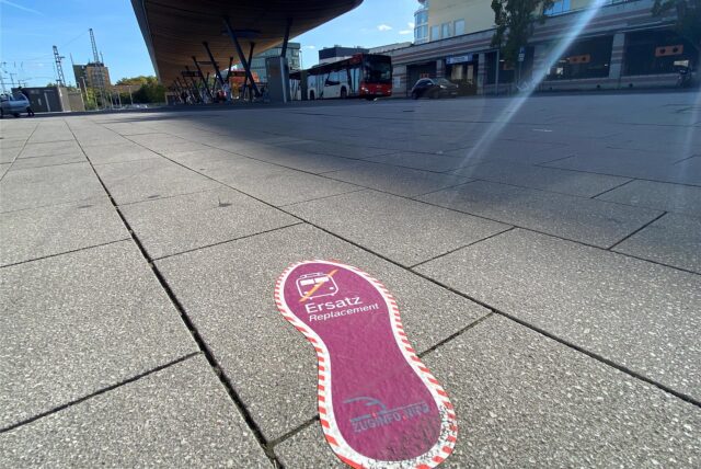 Ein pinker Fußabdruck von Zuginfo.nrw weist auf den Schienenersatzverkehr (SEV) am Bahnhof Unna hin. Im  Hintergrund ist ein Linienbus zu sehen.