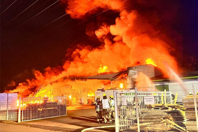 Eine Lagerhalle steht in Vollbrand, Feuerwehrleute stehen mit Abstand davor.