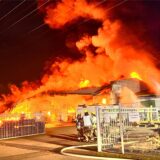 Eine Lagerhalle steht in Vollbrand, Feuerwehrleute stehen mit Abstand davor.