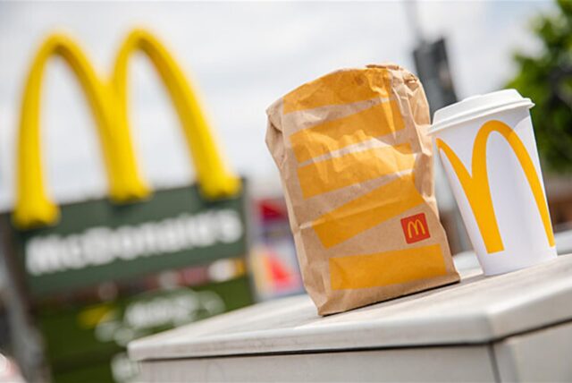 Eine McDonald’s To-Go-Tüte und ein To-Go-Becher stehen vor einer McDonald’s-Filiale.