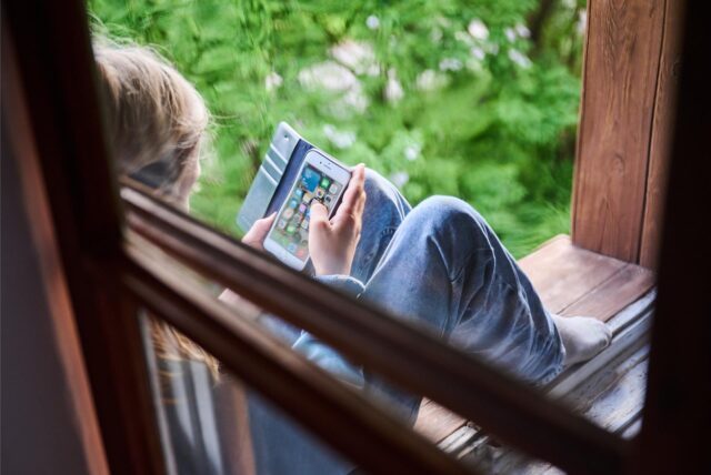 Ein Junge sitzt am Fenster und schaut auf sein Handy.