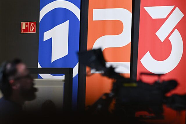 Fahnen mit den Markenlogos von ARD, ZDF und 3Sat sind im Hintergrund einer TV-Kamera zu sehen