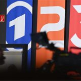 Fahnen mit den Markenlogos von ARD, ZDF und 3Sat sind im Hintergrund einer TV-Kamera zu sehen