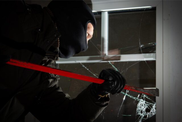 Ein Mann mit Sturmhaube, schwarzer Kleidung und Handschuhen setzt ein Brecheisen an einem Fenster an. Die Scheibe ist gesplittert