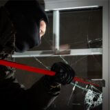 Ein Mann mit Sturmhaube, schwarzer Kleidung und Handschuhen setzt ein Brecheisen an einem Fenster an. Die Scheibe ist gesplittert
