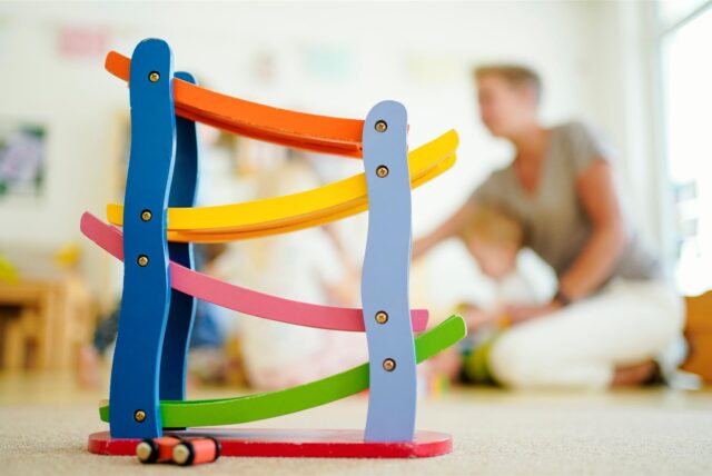Eine bunte Holzbahn steht in einer Kindertagesstätte auf dem Boden. Im Hintergrund spielt eine Frau mit einem Kind