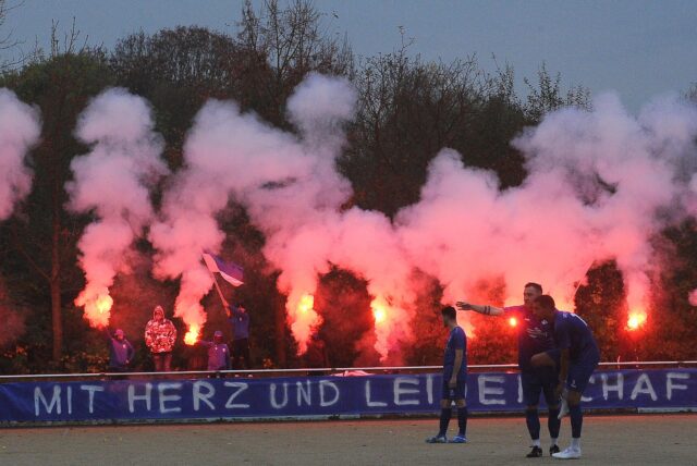 VfL-Schwerte-Fans zünden rote pyrotechnische Fackeln und halten die hinter dem Spruchband „Mit Herz und Leidenschaft“ hoch.