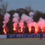 VfL-Schwerte-Fans zünden rote pyrotechnische Fackeln und halten die hinter dem Spruchband „Mit Herz und Leidenschaft“ hoch.