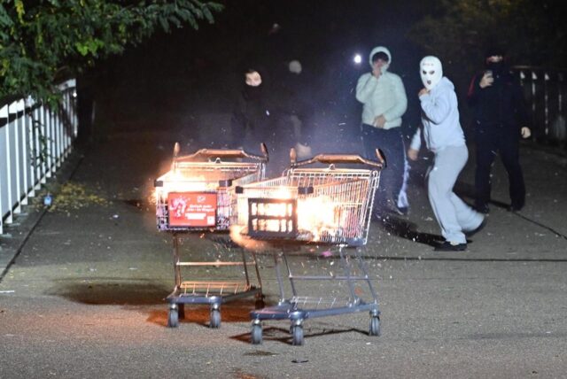 Junge Männer, teils vermummt, schieben brennende Einkaufswagen in Richtung der Einsatzkräfte.