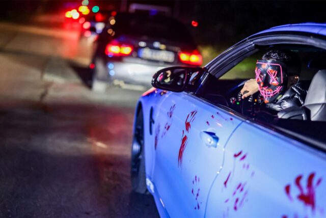 Eine Person mit Maske am Steuer eines weißen Autos, das mit roten Handabdrücken bedeckt ist.