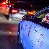 Eine Person mit Maske am Steuer eines weißen Autos, das mit roten Handabdrücken bedeckt ist.