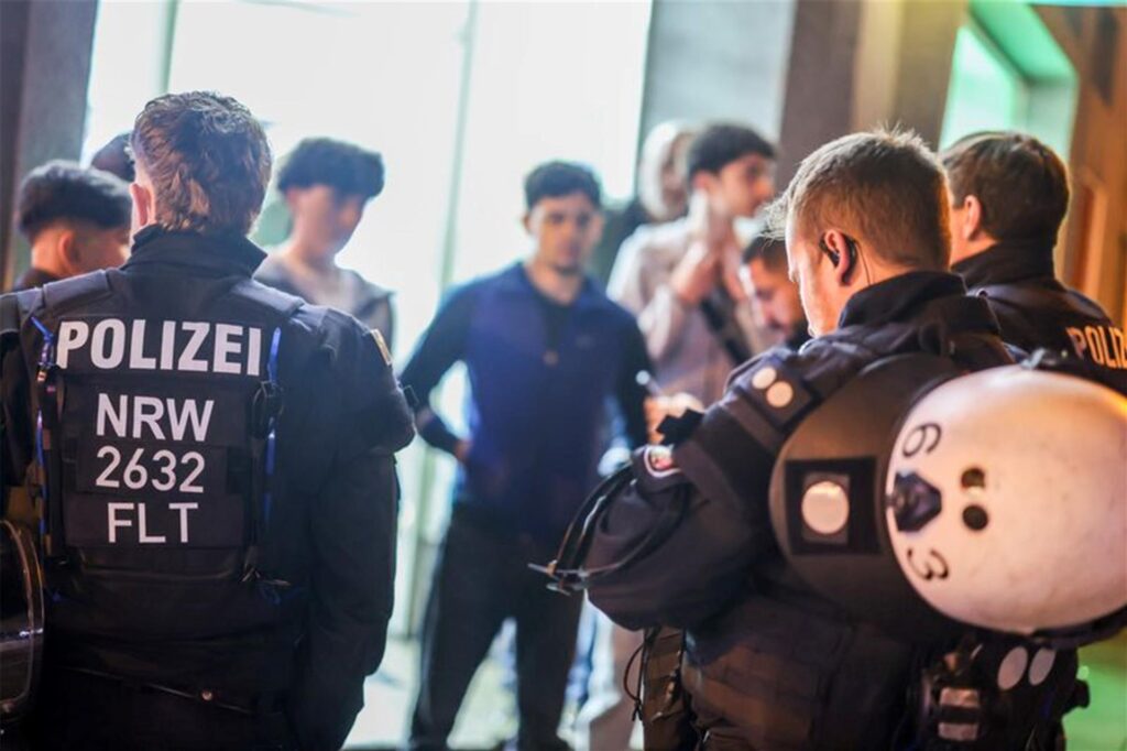 Mehrere Einsatzkräfte der Polizei NRW, im Hintergrund eine Gruppe Männer.