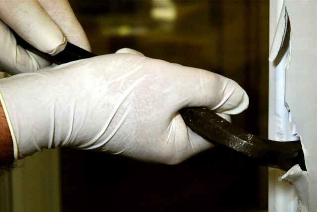 Zwei Hände mit weißen Latexhandschuhen, die mit einem Brecheisen ein Fenster aufhebeln.