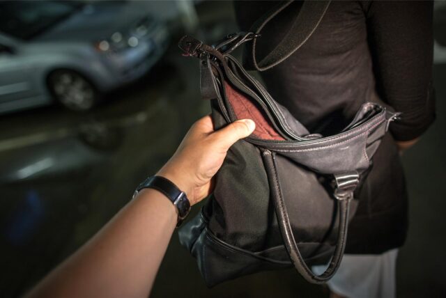 Eine Hand greift nach einer Handtasche, die um die Schulter einer Frau hängt.