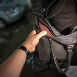 Eine Hand greift nach einer Handtasche, die um die Schulter einer Frau hängt.