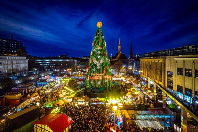 Der große Weihnachtsbaum der Dortmunder Weihnachtsstadt steht inmitten von beleuchteten Ständen und einer großen Menschenmasse. Im Hintergrund sieht man die Skyline der Dortmunder Innenstadt.