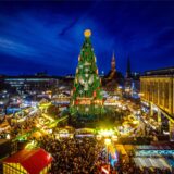 Der große Weihnachtsbaum der Dortmunder Weihnachtsstadt steht inmitten von beleuchteten Ständen und einer großen Menschenmasse. Im Hintergrund sieht man die Skyline der Dortmunder Innenstadt.