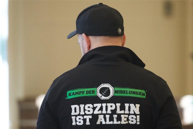 Ein Mann trägt eine Jacke mit der Aufschrift „Kampf der Nibelungen – Disziplin ist alles“, während er in einem Gerichtssaal auf den Beginn des Verfahrens wartet.
