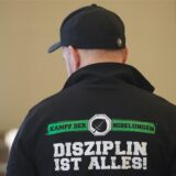 Ein Mann trägt eine Jacke mit der Aufschrift „Kampf der Nibelungen – Disziplin ist alles“, während er in einem Gerichtssaal auf den Beginn des Verfahrens wartet.