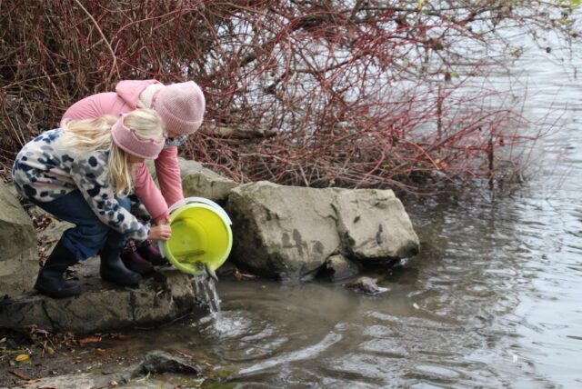 Linn und Matilda halten einen Eimer in die Ruhr, aus dem Fische freigelassen werden.
