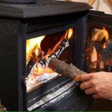 Eine Hand wirft Holz in den brennenden Kamin.