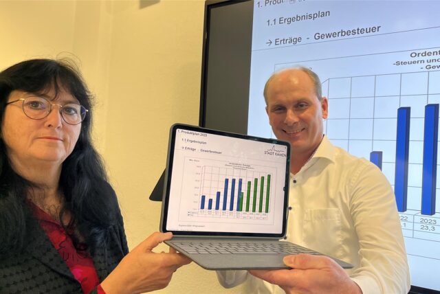 Kamens Bürgermeisterin Elke Kappen und Kämmerer Christian Völkel zeigen auf einem Notebook vor einer Leinwand die aktuellen Zahlen des Kamener Finanzhaushalts.