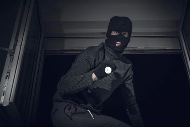 Ein maskierter Einbrecher mit einer Taschenlampe in der Hand steigt durch ein Fenster. (gestellte Szene)