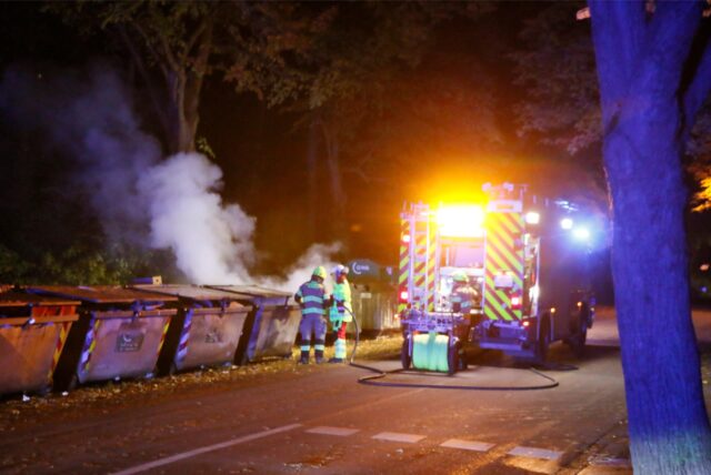 Feuerwehrleute löschen ein Feuer in einem Müllcontainer in Kamen. Aus dem grauen Papiersammler quillt Rauch.