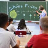 Eine Lehrerin schreibt „Schöne Ferien“ mit Kreide an eine Schultafel und die Schüler sitzen an den Tischen.