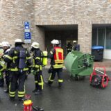 Vor dem Unnaer Rathaus stehen während eines Einsatzes 2019 Feuerwehrleute, teils mit Atemschutzgeräten, und eine Polizistin. Das Bild zeigt auch einen durch Feuer zerstörten Müllcontainer und ein Lüftungsgerät.
