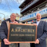 Zwei Männer halten gemeinsam ein Schild und stehen vor einem Elektrofachgeschäft.