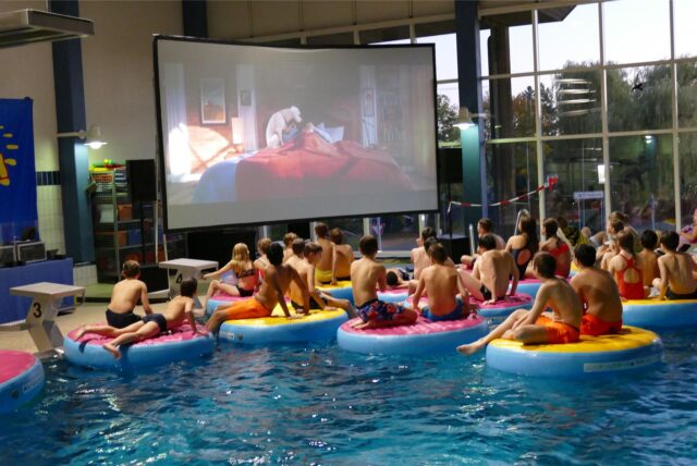 Jugendliche in Badesachen sitzen auf schwimmenden Sesseln im Hallenbad in Bergkamen und schauen auf einer Leinwand am Beckenrand einen Animationsfilm.
