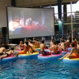 Jugendliche in Badesachen sitzen auf schwimmenden Sesseln im Hallenbad in Bergkamen und schauen auf einer Leinwand am Beckenrand einen Animationsfilm.